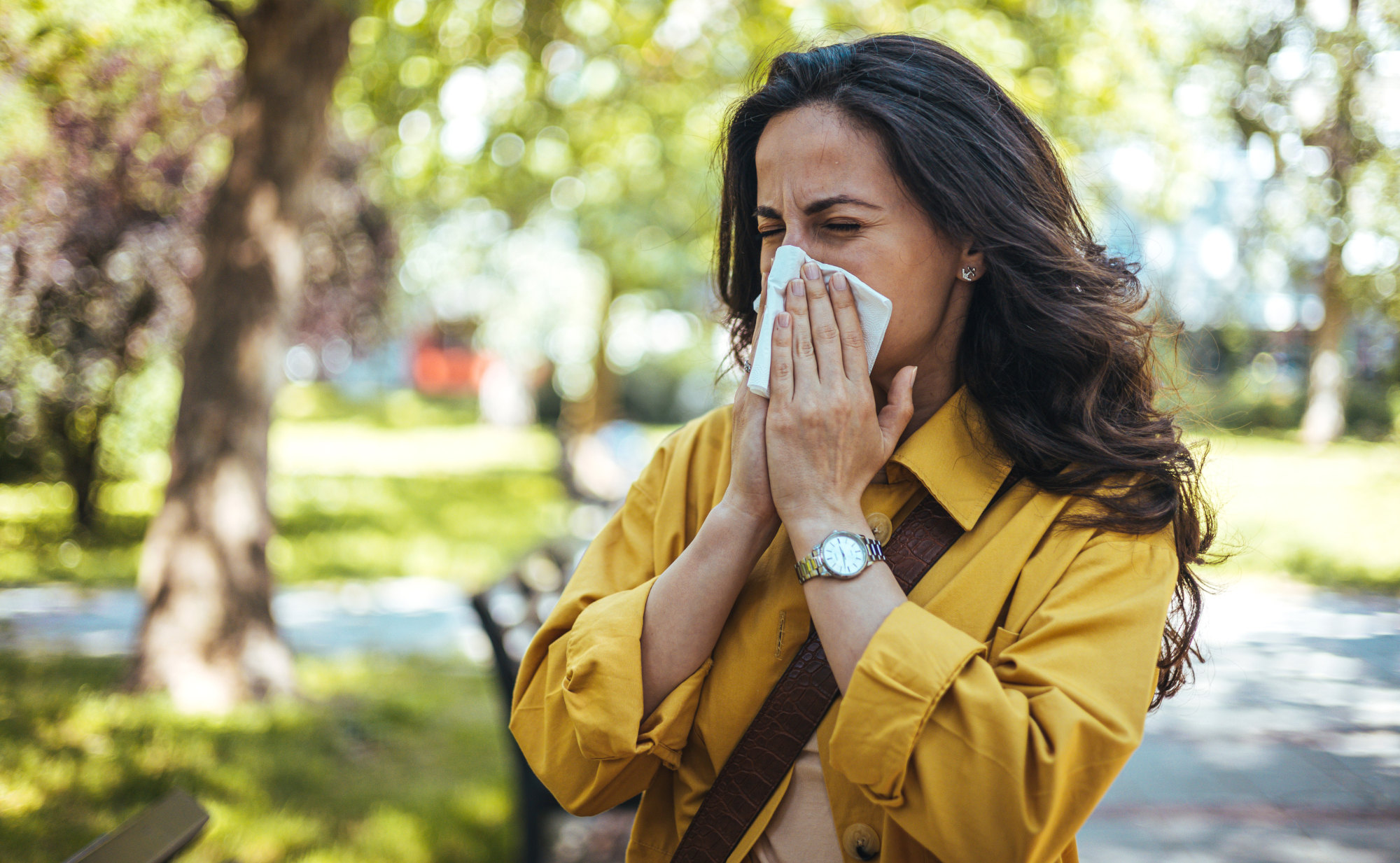 Eine Frau mit langen, dunklen Haaren niest oder putzt sich die Nase mit einem Taschentuch in einem sonnigen Park. Ein Symbolfoto von LIFE LIGHT zum Thema Allergien im Zusammenhang mit dem Immunsystem.