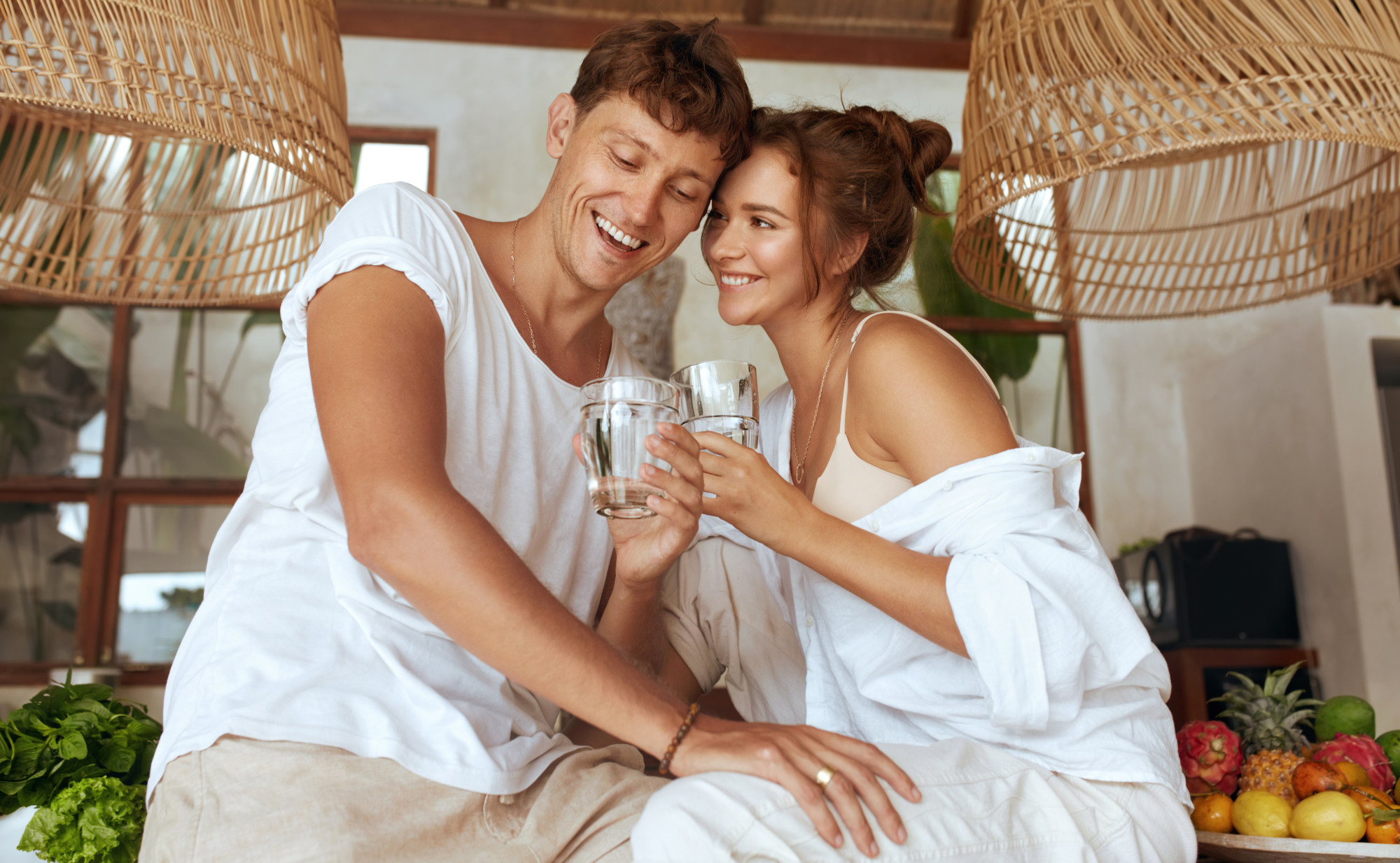 Ein Mann und eine Frau stoßen mit Wassergläsern an und lächeln sich in einer Küche an. Ein Symbolfoto für Wohlbefinden durch Entgiftung und Detox.