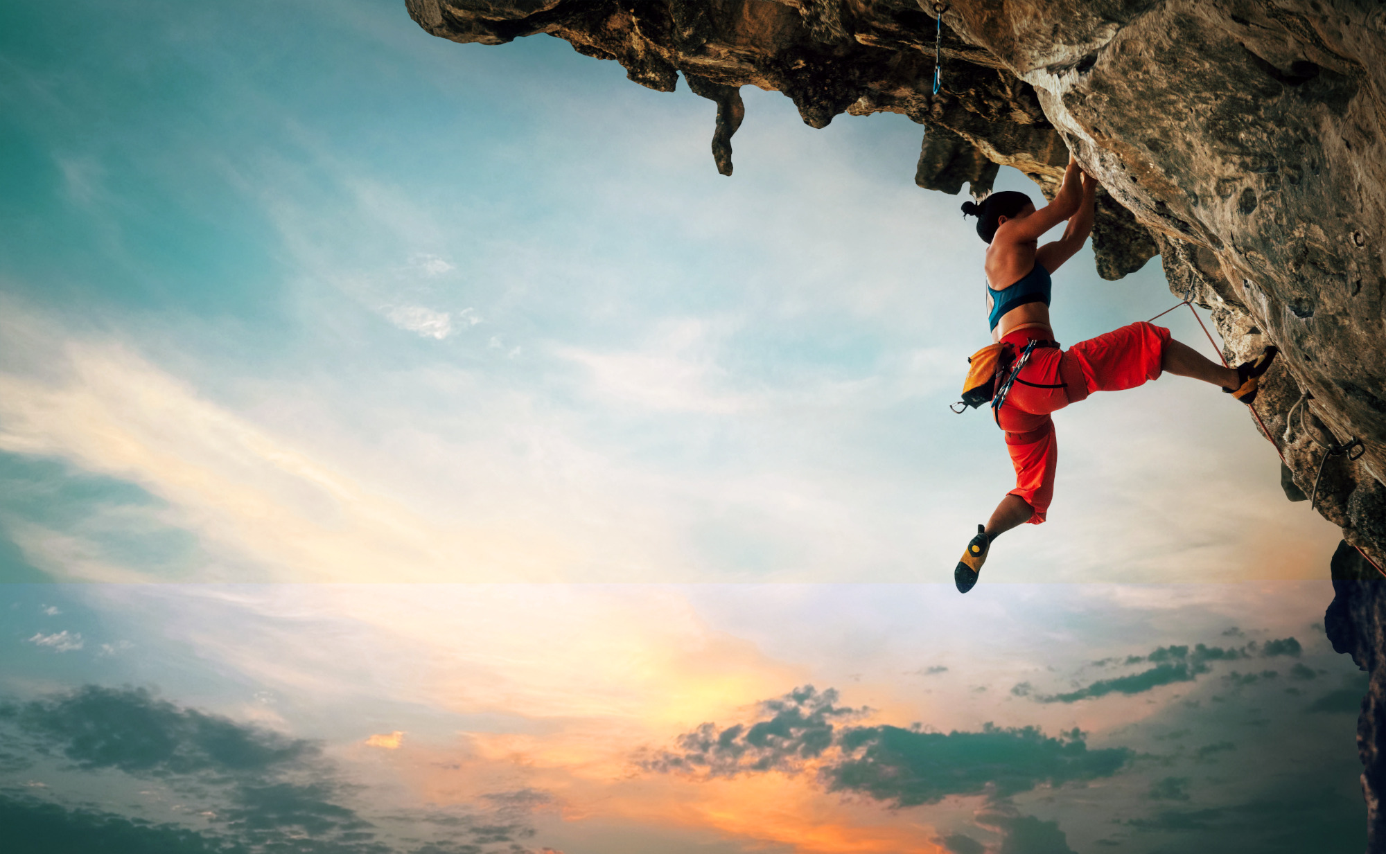 Eine Frau klettert an einer überhängenden Felswand, während die Sonne untergeht. Ein Symbolfoto für Kraft und Energie anhand ausgewählter Mikronährstoffe.