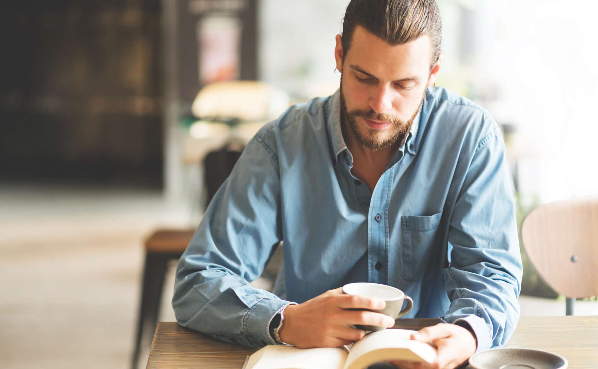 Ein Mann mit Bart sitzt in einem Café, liest ein Buch und hält eine Tasse in der Hand. Ein Symbolfoto für Nährstoffe als Energiequelle fürs Gehirn.