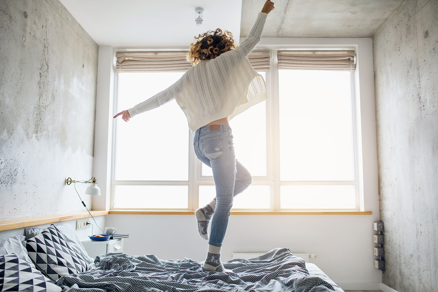 Eine Frau mit lockigen Haaren springt in einem Schlafzimmer auf einem Bett, die Arme ausgestreckt und den Rücken zum Betrachter. Ein Symbolfoto für Lebensenergie durch gestärkte Mitochondrien.