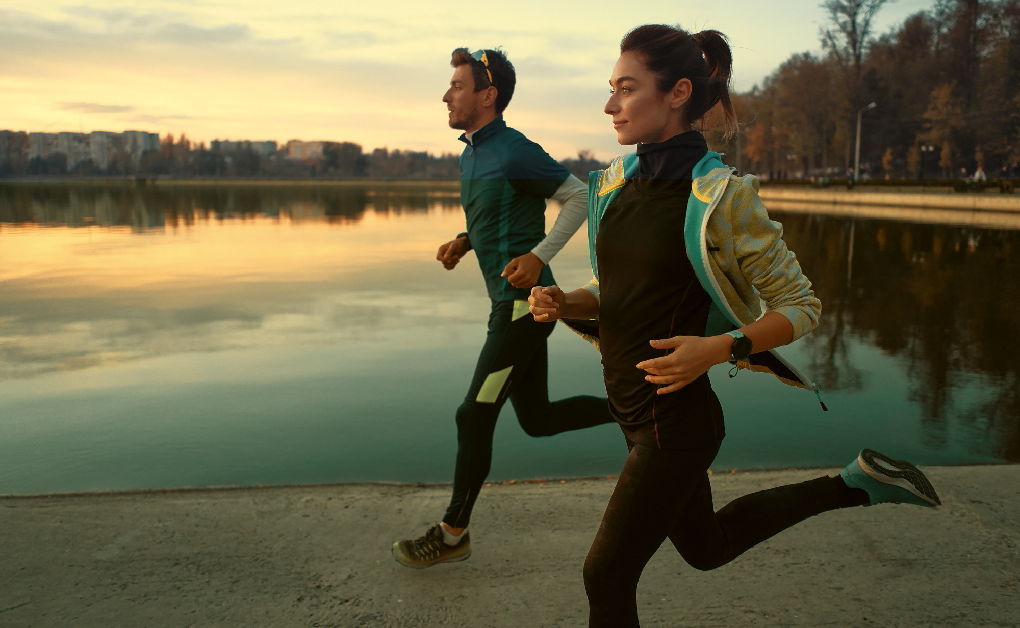 Ein Mann und eine Frau joggen am Ufer eines Sees bei Sonnenuntergang. Die Frau im Vordergrund lächelt. Ein Symbolfoto für Beweglichkeit und gesunde Gelenke.