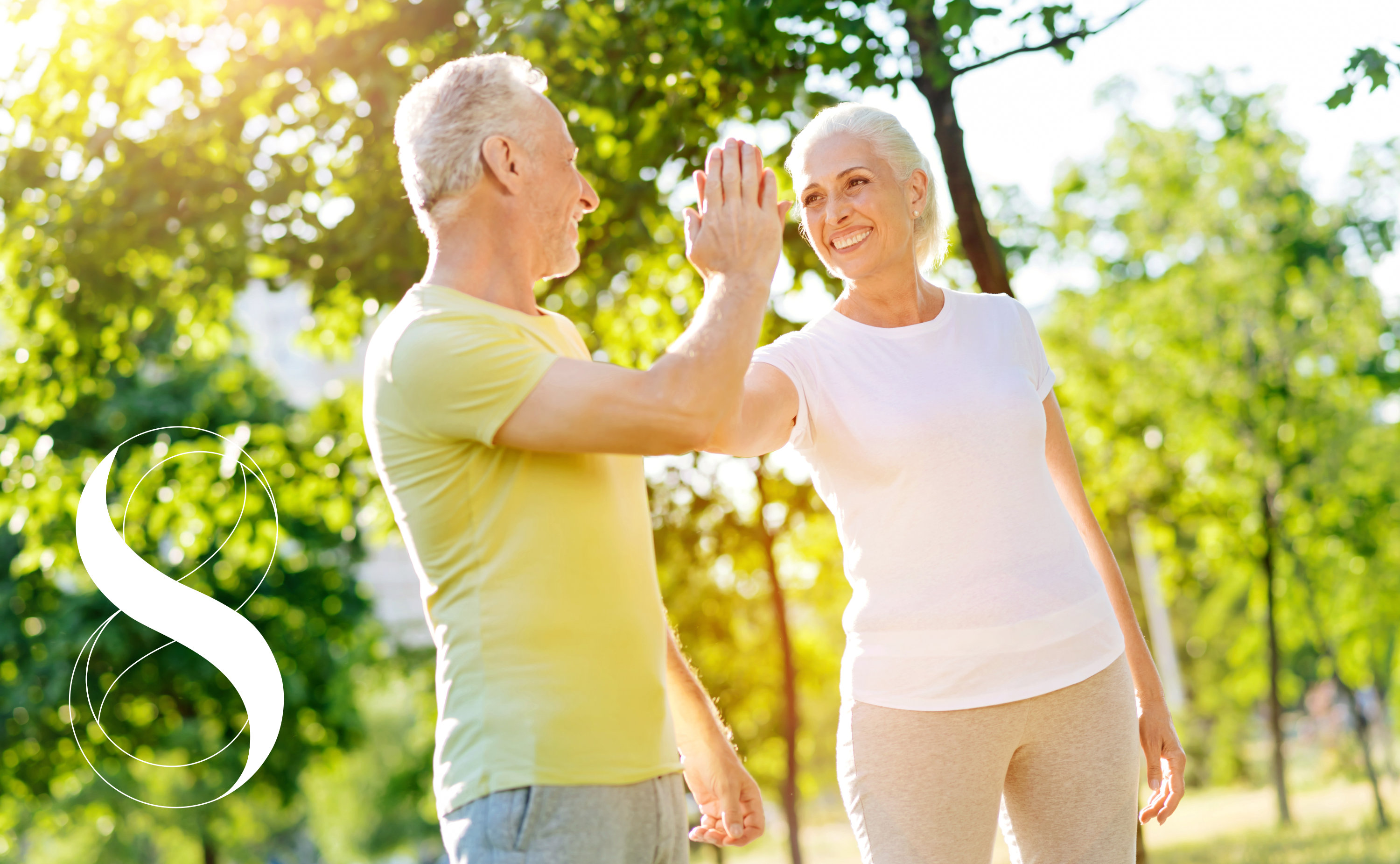 Ein Mann und eine Frau in höherem Alter, beide sportlich gekleidet in einem sonnigen Park, die sich gegenseitig lachend abklatschen. Sie symbolisieren Vitalität durch gesunde Gelenke.