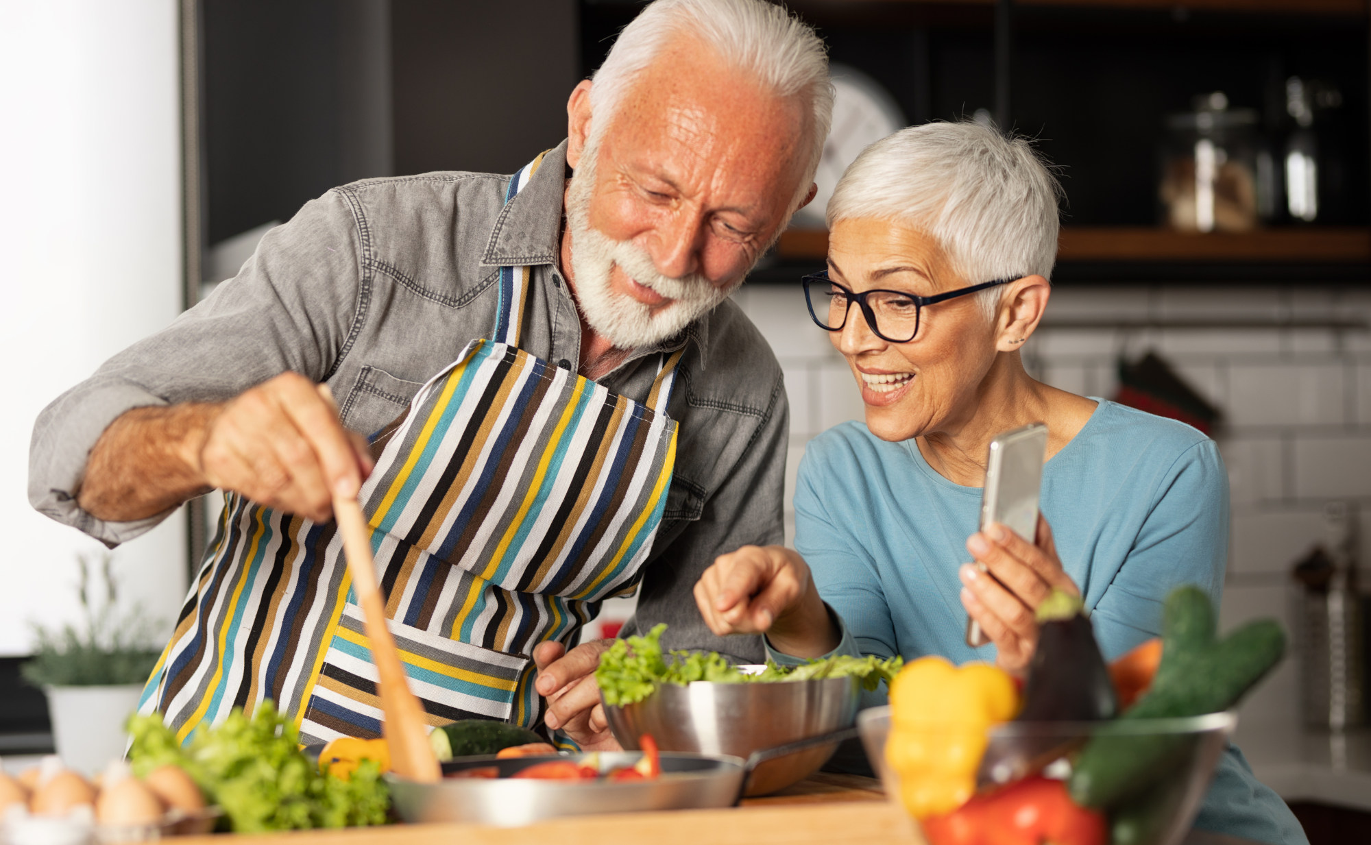 Ein Mann und eine Frau, beide mit mit weißen Haaren kochen zusammen in einer Küche und schauen freudig auf das Essen. Ein Symbolfoto für natürliches Gewichtsmanagement mit gesunden Lebensmitteln.