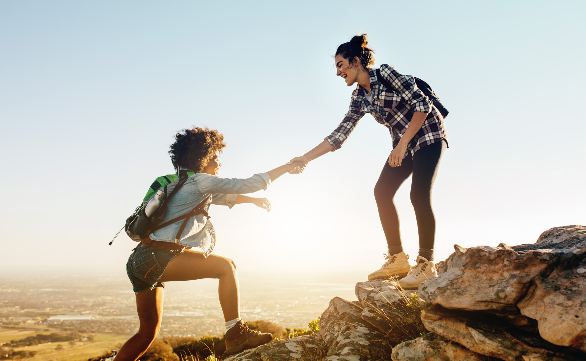 Zwei Wanderinnen am Berg: Eine junge Frau steht auf einem Felsen und reicht einer anderen jungen Frau die Hand, die ihr auf allen Vieren den Berg entgegen klettert. Ein Symbolfoto von LIFE LIGHT für das Verlassen von Komfortzonen.