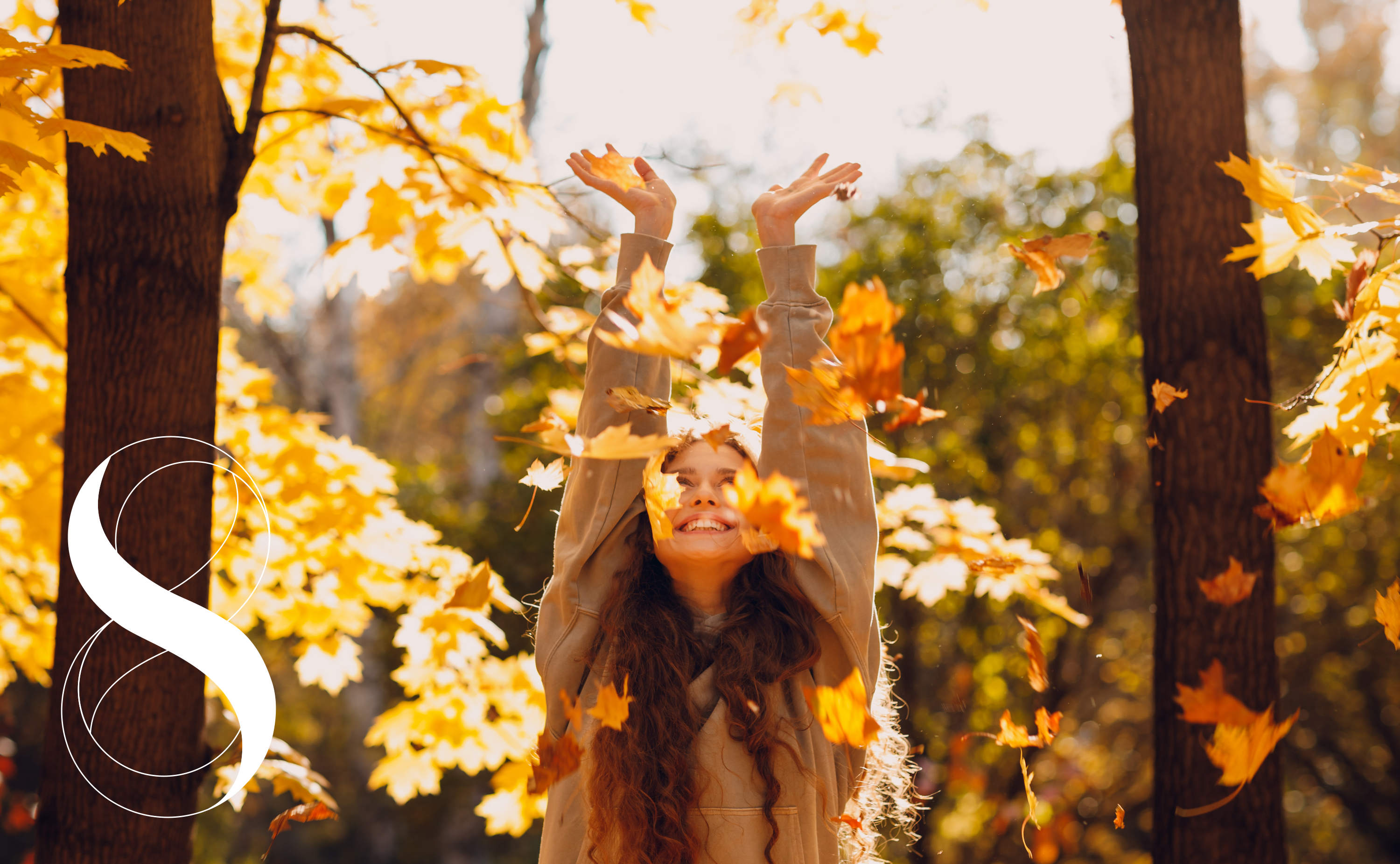 Mädchen mit langen, welligen Haaren in beigem Pullover, das im Herbstwald steht. Sie lacht und streckt ihre Arme nach oben, während goldene und orangefarbene Blätter herabfallen. Ein Symbolfoto für Krafttanken im Herbst mit einem Magnesium-Komplex von LIFE LIGHT.