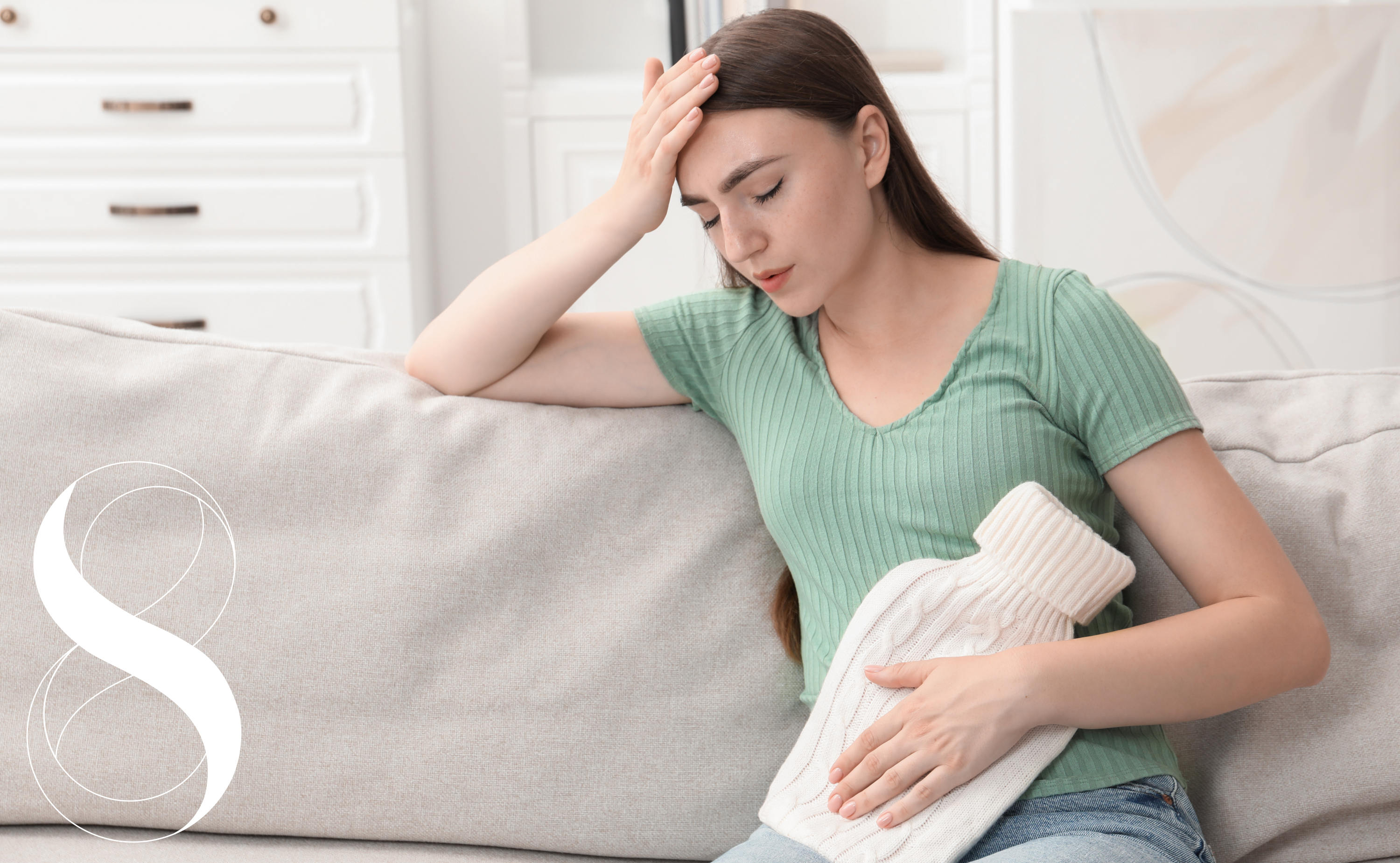 Eine junge Frau sitzt mit schmerzverzerrtem Gesicht auf einem hellgrauen Sofa. Sie hält sich eine weiße Wärmeflasche gegen ihren Unterbauch und stützt ihren Kopf erschöpft mit der Hand an der Stirn ab. ein Symbolbild von LIFE LIGHT für PMS.