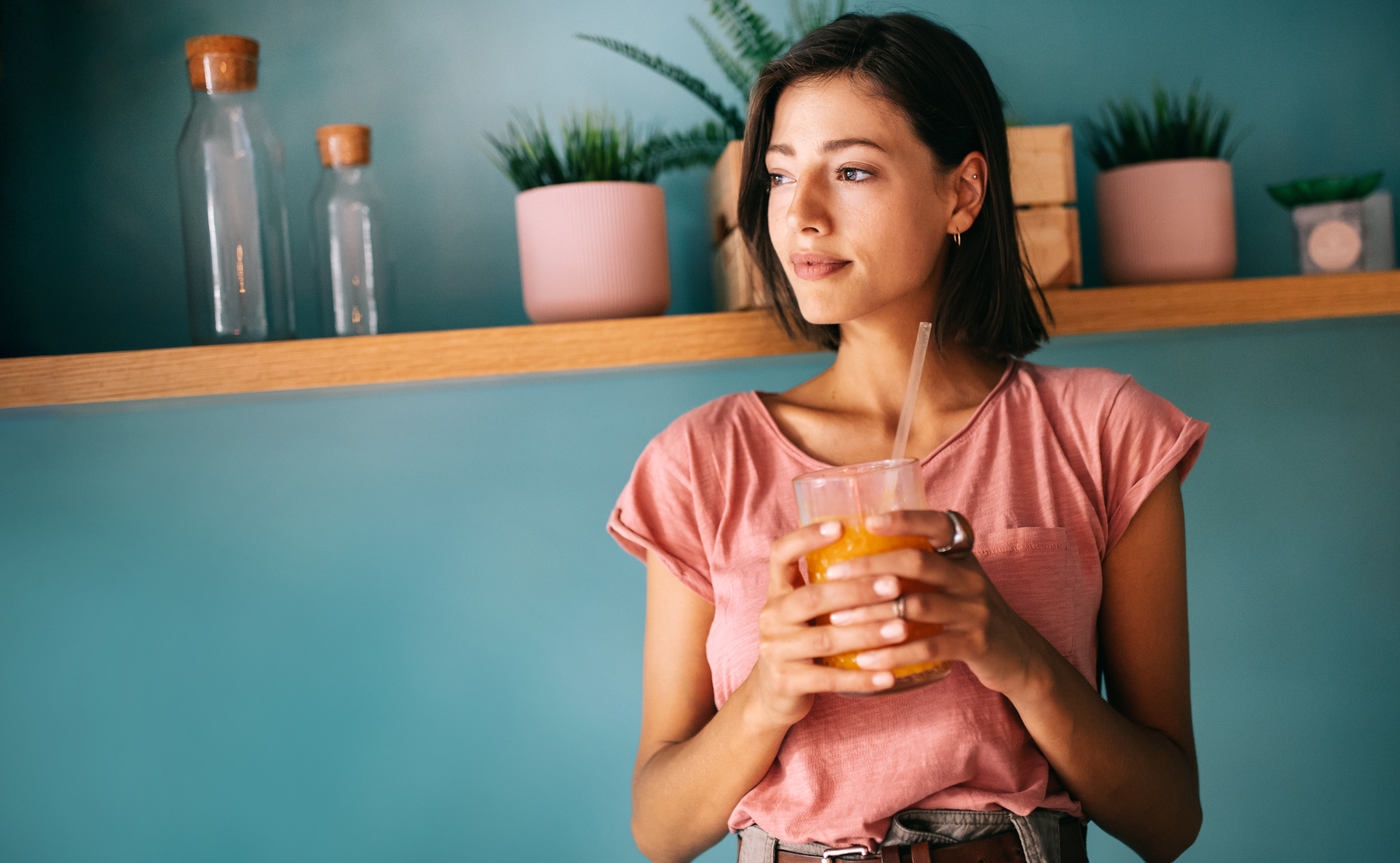 Eine junge Frau in einem Café hält ein Glas mit gelbem Saft und einem Strohhalm in den Händen und blickt zur Seite. Ein Symbolfoto für den Genuss gesunder Pflanzensäfte.