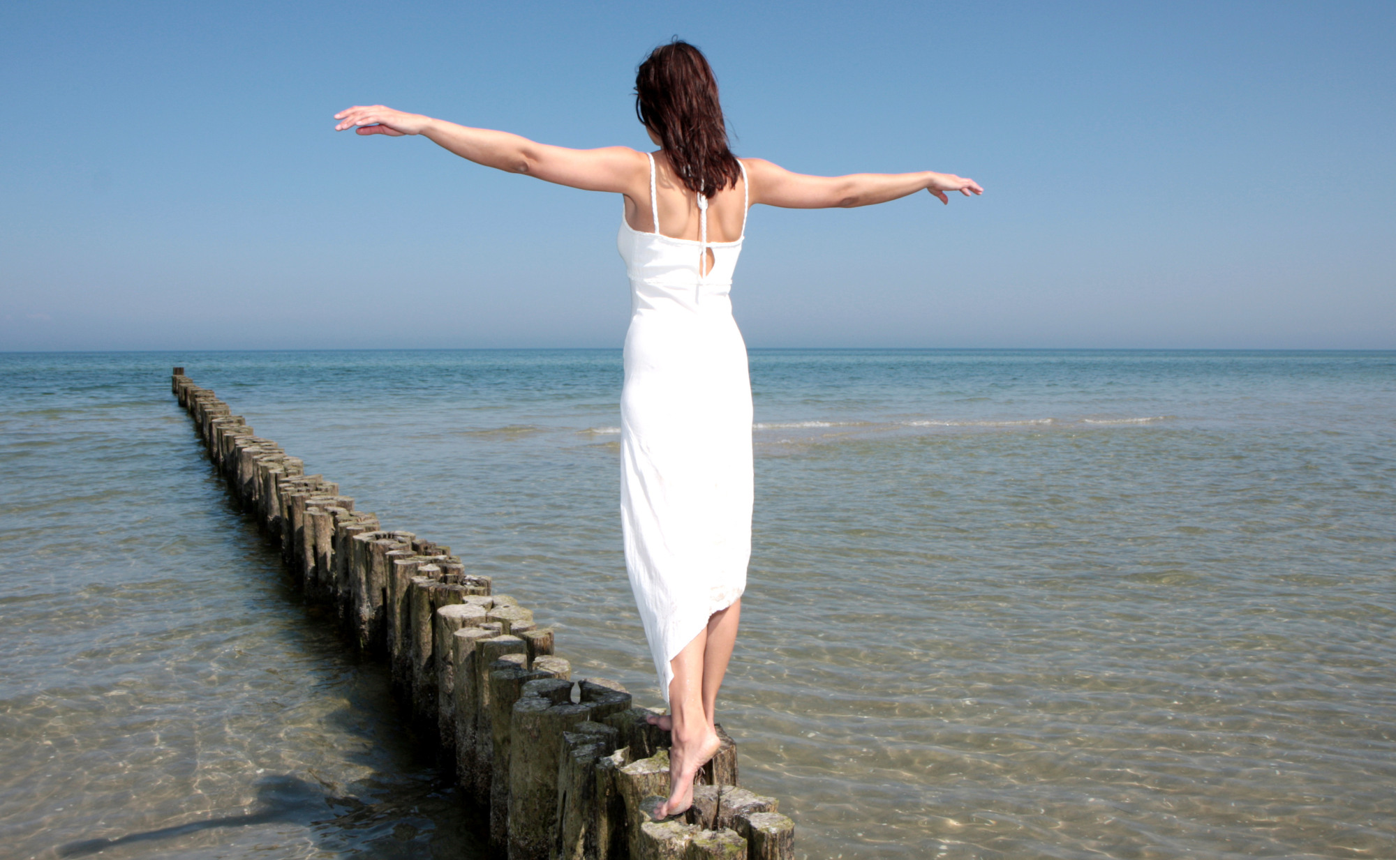 Eine Frau in einem weißen Kleid balanciert mit ausgebreiteten Armen auf einigen Bolzbalken im flachen Meer. Ein Symbolfoto von LIFE LIGHT für Balance - auch im Säure-Basen-Haushalt.