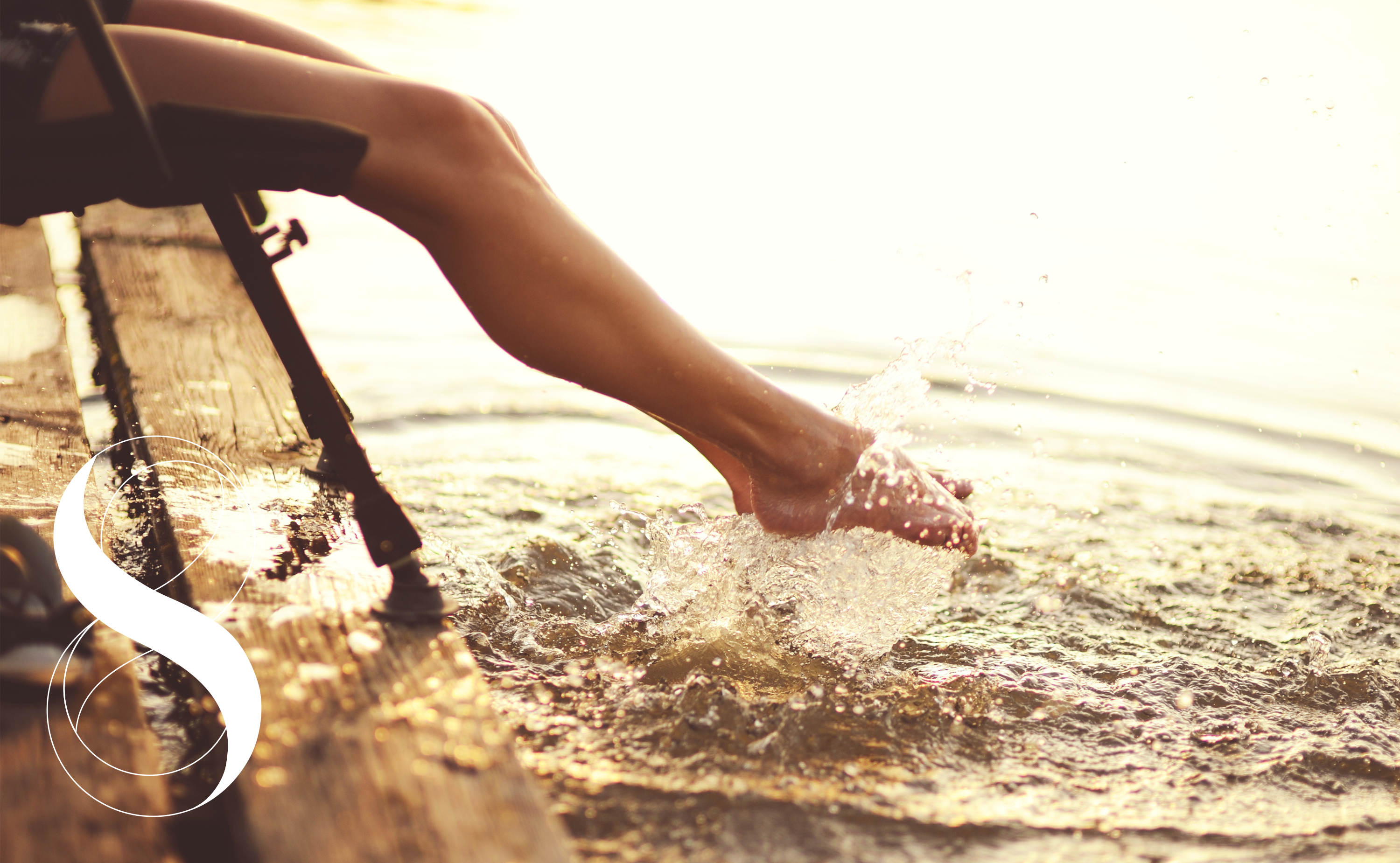 Eine Frau sitzt auf einem Steg und verspritzt mit ihren Beinen das Wasser unter ihr, während das Sonnenlicht die Szene in warmes Licht taucht. Ein Symbolbild von Life Light für gesunde, gekühlte Beine ohne Ödeme.
